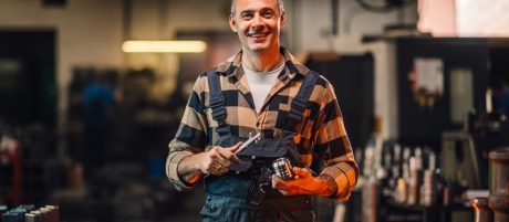 A picture of a metallurgy mechanic holding a tool and a cylinder machine element, smiling at the camera. Blurred background of manufacture facility with various metal shapes and machines.